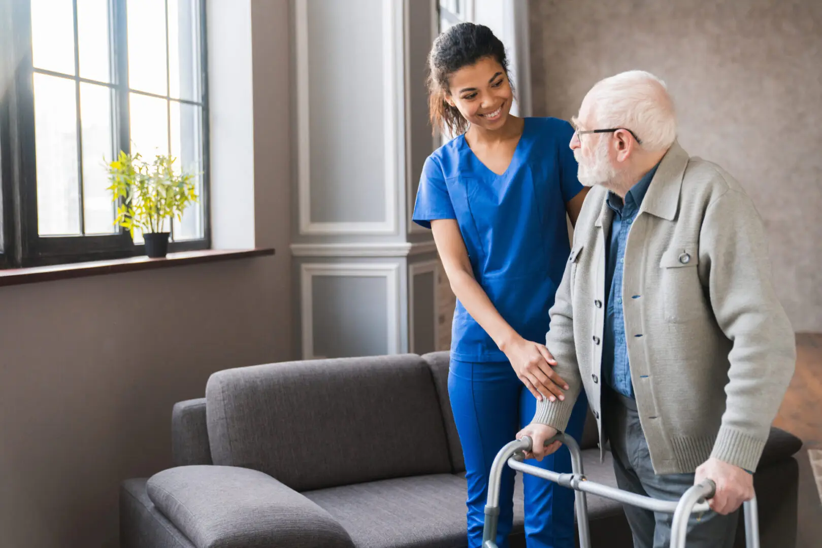Portrait of an african young nurse helping old elderly disable man grandfather to walk using walker equipment in the bedroom. Senior patient of nursing home moving with walking frame and nurse support Caregiver assisting elderly man with walker.